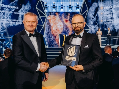 Een medium-long shot van twee mannen in formele avondkleding die elkaar de hand schudden. De man rechts, Maciej Góralski, houdt een ingelijste onderscheiding vast met een gouden embleem, waarop te lezen staat dat het de prijs voor „Ondernemer van het Jaar 2022“ betreft. Op de wazige achtergrond is een podium te zien met abstracte geometrische structuren en dynamische blauwe en roodoranje grafische patronen. In de slecht verlichte evenementenzaal zitten verschillende gasten aan tafels.