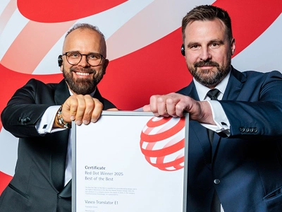 Twee mannen in formele zakelijke kleding – de man links in een smoking met een bril, de man rechts in een donkerblauw pak – houden samen met beide handen een ingelijst certificaat vast. Op het certificaat staat duidelijk vermeld: „Red Dot-winnaar 2025 Best of the Best“, toegekend aan „Vasco Translator E1“. Op de achtergrond is een grootschalige, dynamische grafische achtergrond te zien met abstracte rode en witte golfpatronen.