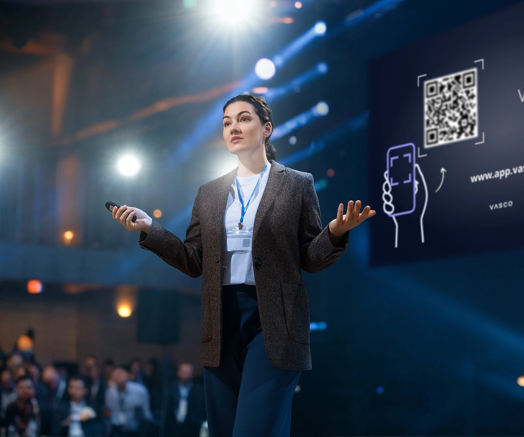 Een vrouw staat op een helder verlicht conferentiepodium en houdt een presentatie voor het publiek. Ze draagt een bruine blazer, een wit T-shirt en een conferentiepas aan een koord. In haar rechterhand houdt ze een afstandsbediening voor de presentatie vast en met haar linkerhand maakt ze gebaren. Achter haar is op een groot scherm een opvallende QR-code voor de dienst 'Vasco Assistance' te zien, samen met een kleine afbeelding van een smartphone die deze code scant. Het publiek op de voorgrond is wazig.