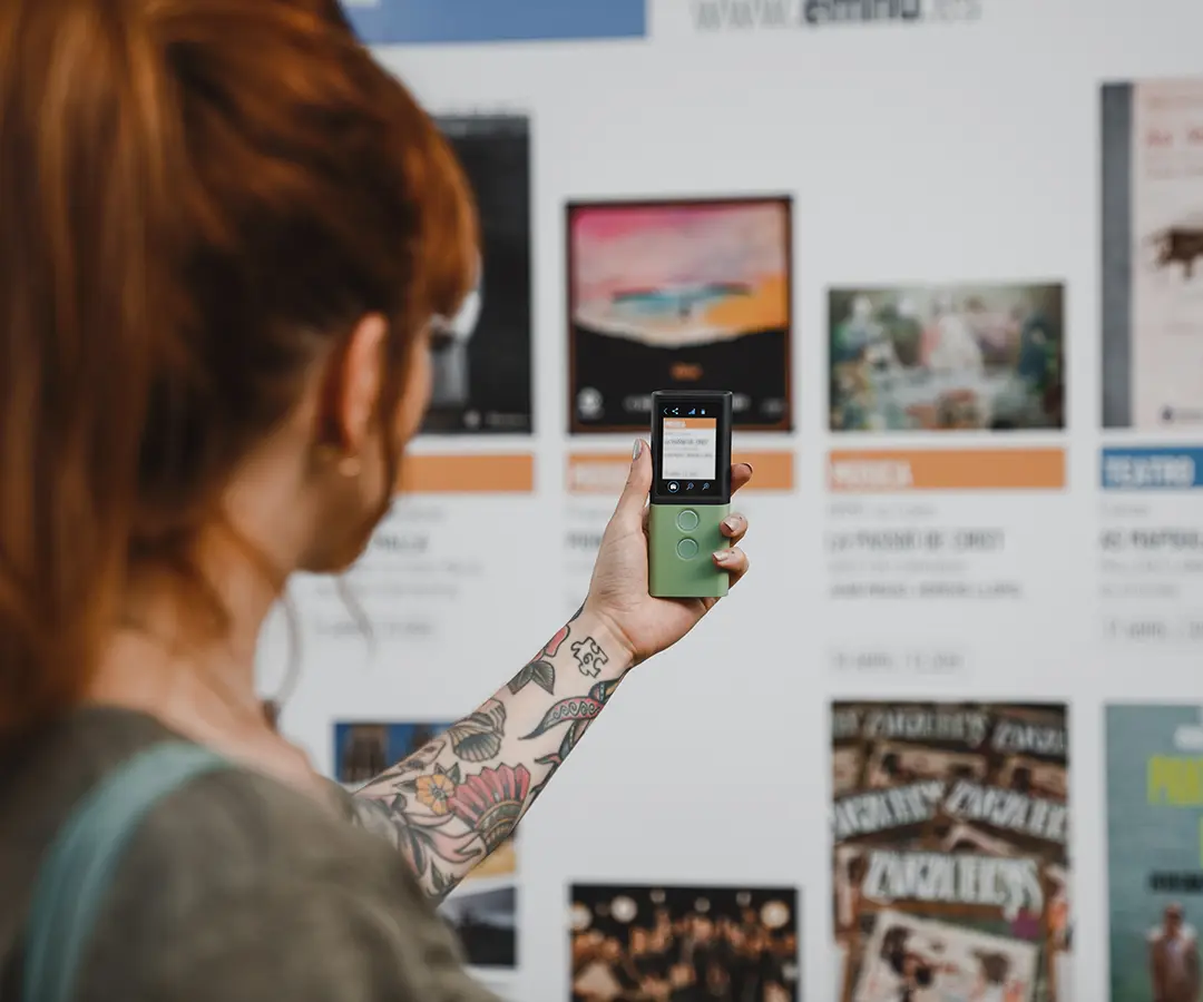 Iemand met rood haar en een bloementatoeage op zijn arm houdt een groen digitaal apparaat omhoog en richt het op een displaybord met verschillende posters en informatie over evenementen.
