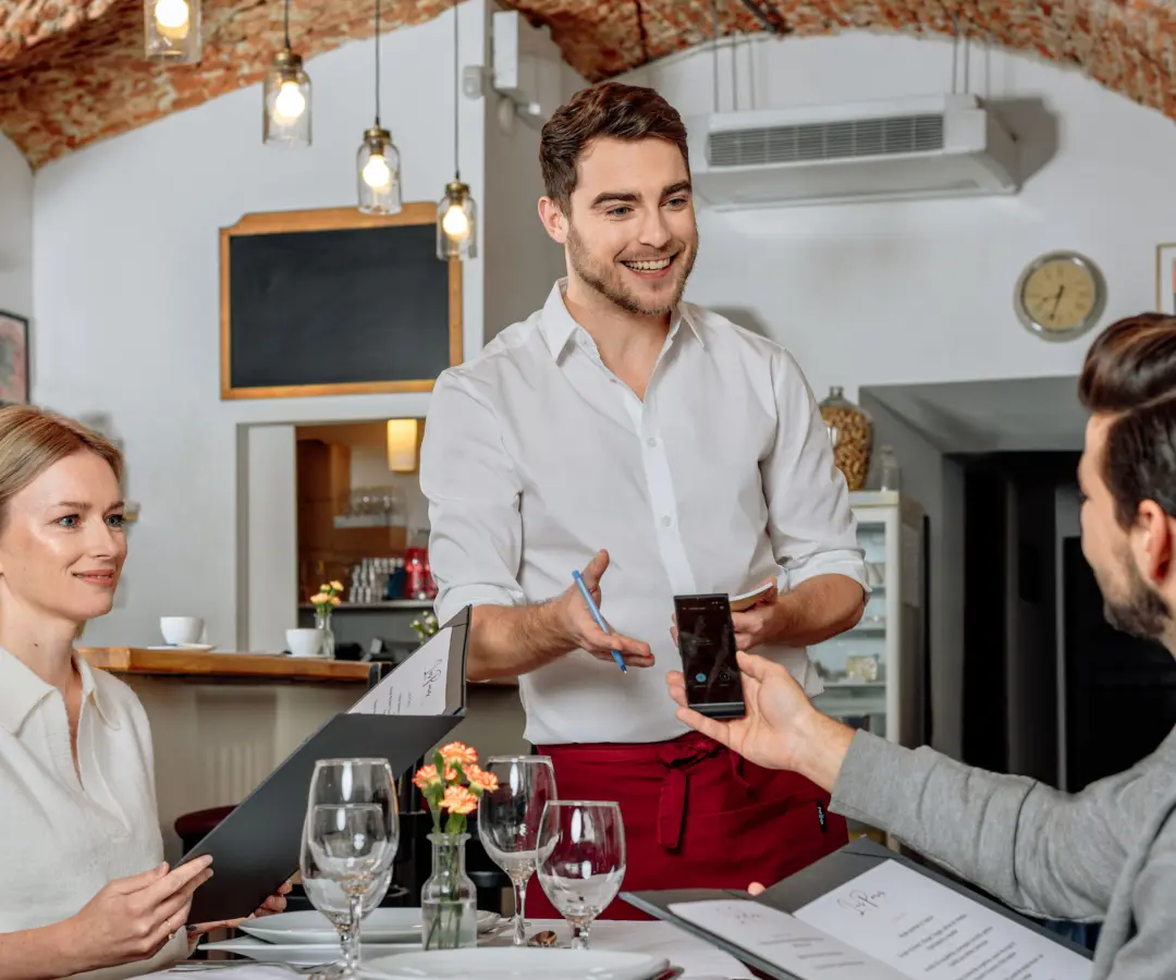 Een glimlachende ober staat bij een restauranttafel, terwijl een zittende mannelijke klant een vertaler in zijn hand houdt. Een vrouwelijke klant zit vlakbij en houdt een menukaart vast. De tafel is gedekt met glazen, borden en een kleine bloemenvaas.