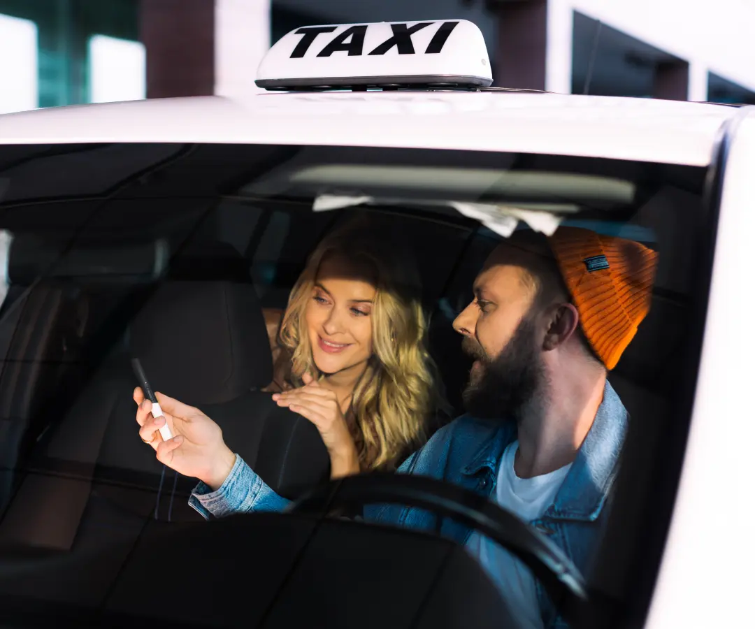 Een man zit op de bestuurdersstoel van een taxi en laat iets zien aan een vrouw op de achterbank op een vertaler die hij in zijn hand houdt. De vrouw glimlacht geïnteresseerd. Op het dak van de auto is een taxibord zichtbaar.