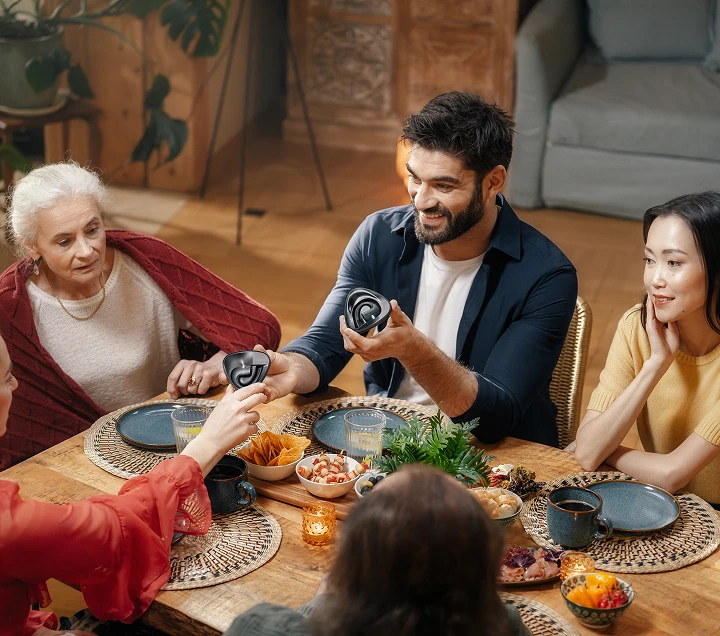 Mensen uit verschillende landen zaten rond een tafel, genoten van een maaltijd en voerden levendige gesprekken, waardoor er een warme en uitnodigende sfeer ontstond terwijl ze de oortjes van de Vasco Translator E1 aan elkaar doorgaven.