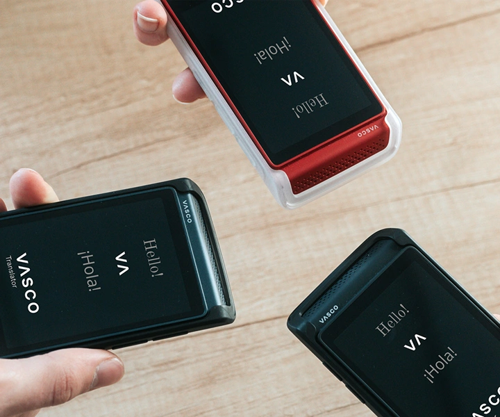 Drie mensen met een Vasco Q1 in de hand staan bij elkaar; op de schermen is het startscherm te zien met het Vasco logo en de woorden ‘Hello!’ en ‘¡Hola!’. De apparaten worden gepresenteerd in siliconenhoesjes in drie kleuren: Solid Black, Dark Caramel en Milky White.