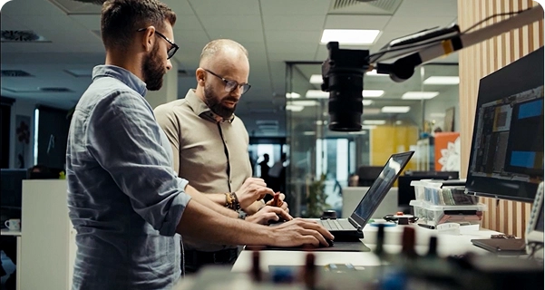 Twee mannen met baarden en een bril werken samen aan een sta-bureau in een modern kantoor of laboratorium. De ene man, in een lichtblauw overhemd, werkt op een laptop, terwijl de andere man, in een beige overhemd, naast hem staat en naar een klein elektronisch onderdeel in zijn hand wijst. Op het bureau staan een groot beeldscherm, diverse elektronische apparaten en een camera die op een verstelbare arm is bevestigd. Op de achtergrond is een lichte, open werkruimte met glazen scheidingswanden te zien. 