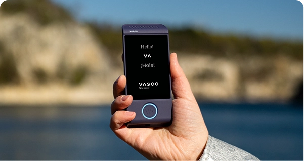 Een close-up van een hand die een paars elektronisch vertaalapparaat van Vasco vasthoudt, tegen een heldere buitenachtergrond. Op het scherm staan de begroetingen 