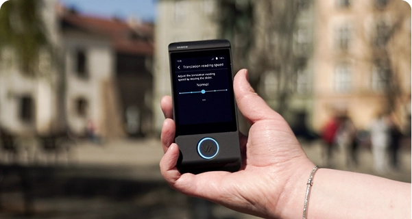 Een close-up van een hand die een zwarte elektronische vertaler van Vasco vasthoudt, tegen een wazige buitenachtergrond van een stadsplein met historische gebouwen. Op het scherm van het apparaat zijn de instellingen voor de „leessnelheid van de vertaling“ te zien, met een schuifbalk die op „Normaal“ staat en een opmerking die luidt: „Pas de leessnelheid van de vertaling aan door de schuifbalk te verplaatsen.“ De persoon die het apparaat vasthoudt, draagt een zilveren armband. Het felle daglicht laat de interface van het scherm goed uitkomen, waardoor de instellingen duidelijk zichtbaar zijn. 