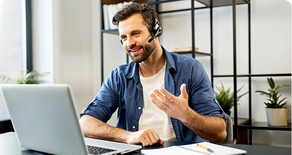 Een vriendelijk ogende man met een baard en donker haar zit aan een bureau, met een professionele headset met microfoon op. Hij glimlacht en gebaart met één hand terwijl hij naar het scherm van een laptop kijkt; het lijkt alsof hij midden in een videogesprek zit of technische ondersteuning biedt. Hij draagt een blauw overhemd over een wit T-shirt. Op het bureau voor hem liggen een laptop, een notitieboekje en een gele pen. De achtergrond is een lichte, moderne kantoorruimte met een minimalistische zwarte metalen plank en groene potplanten. 