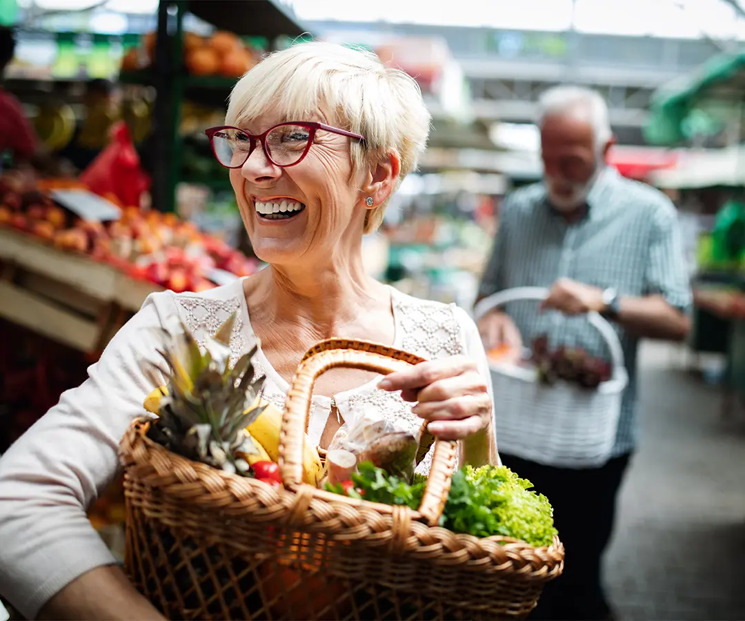 Een glimlachende oudere vrouw met kort blond haar en een rode bril, die op een openluchtmarkt een rieten mand vol verse groenten en fruit bij zich heeft. Op de wazige achtergrond is nog iemand te zien die tussen de kraampjes met groenten en fruit aan het winkelen is.
