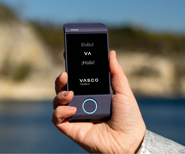 A close-up of a hand holding a purple Vasco Translator M4 electronic translator device against a bright outdoor background. The screen displays the greetings "Hello!", "¡Hola!", and the "VASCO Translator" logo.